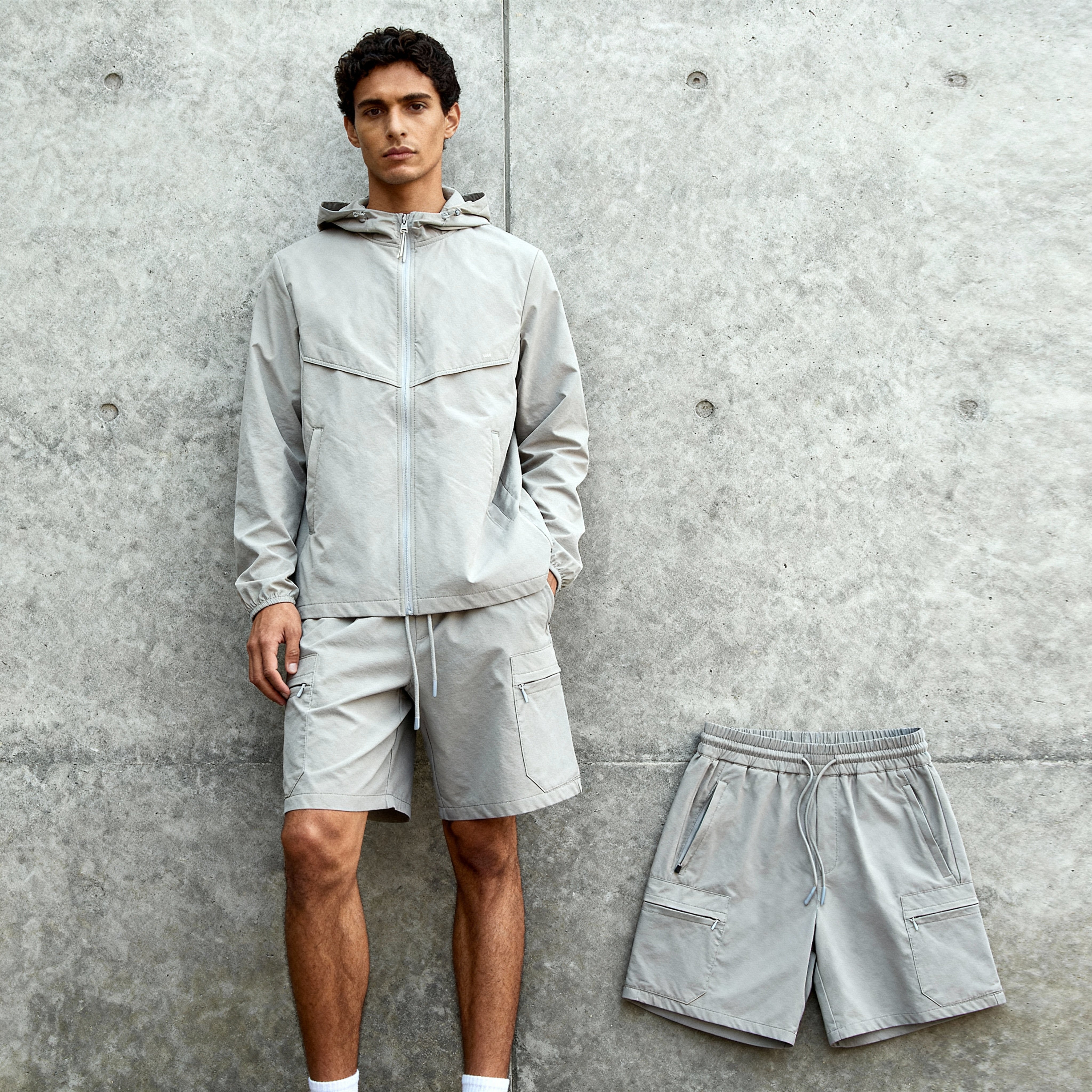 Man wearing a light gray jacket and shorts against a concrete wall, with another pair of gray shorts on the ground.