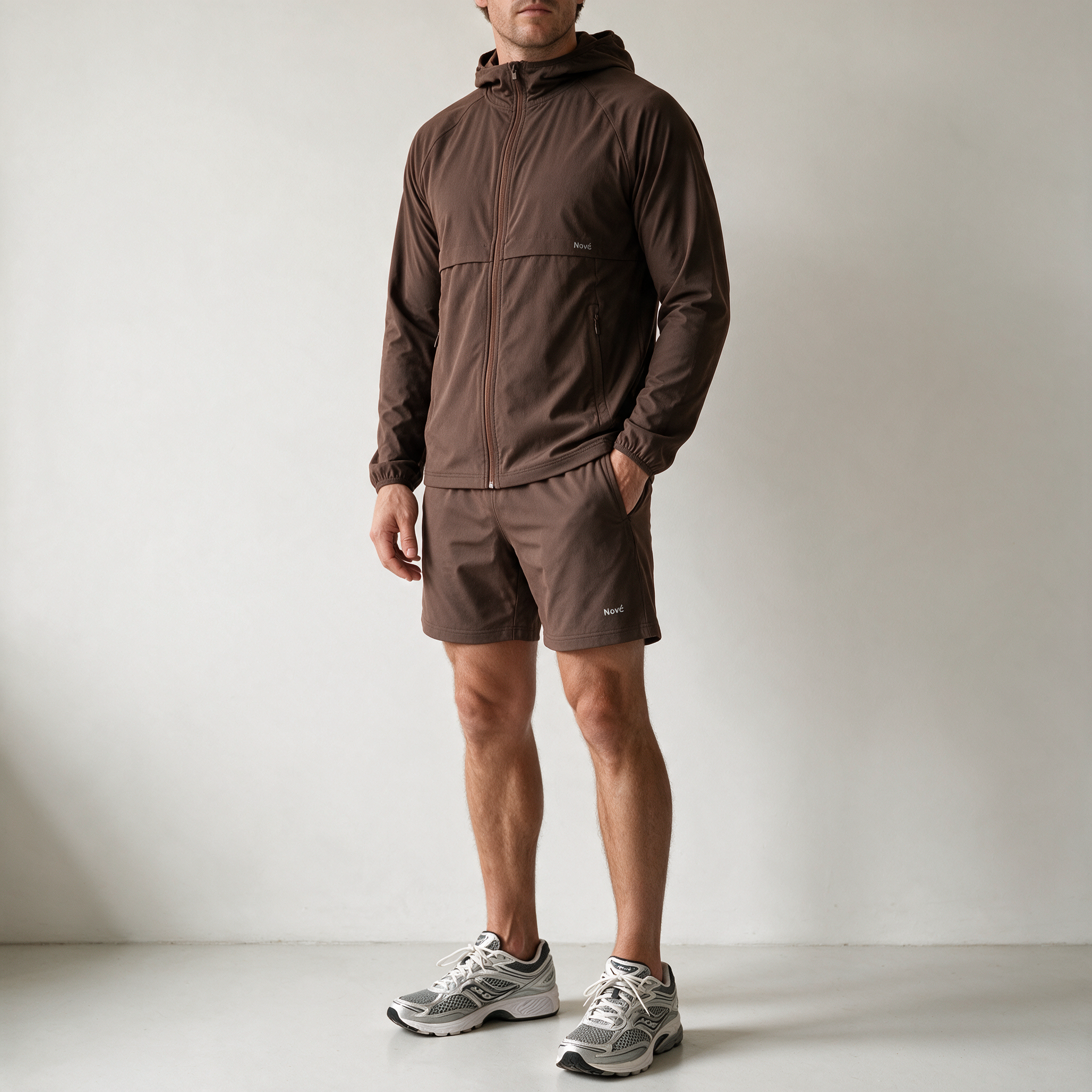 A man wearing a brown hooded zip-up jacket and matching brown shorts, paired with gray and white running shoes, standing against a plain light gray wall.