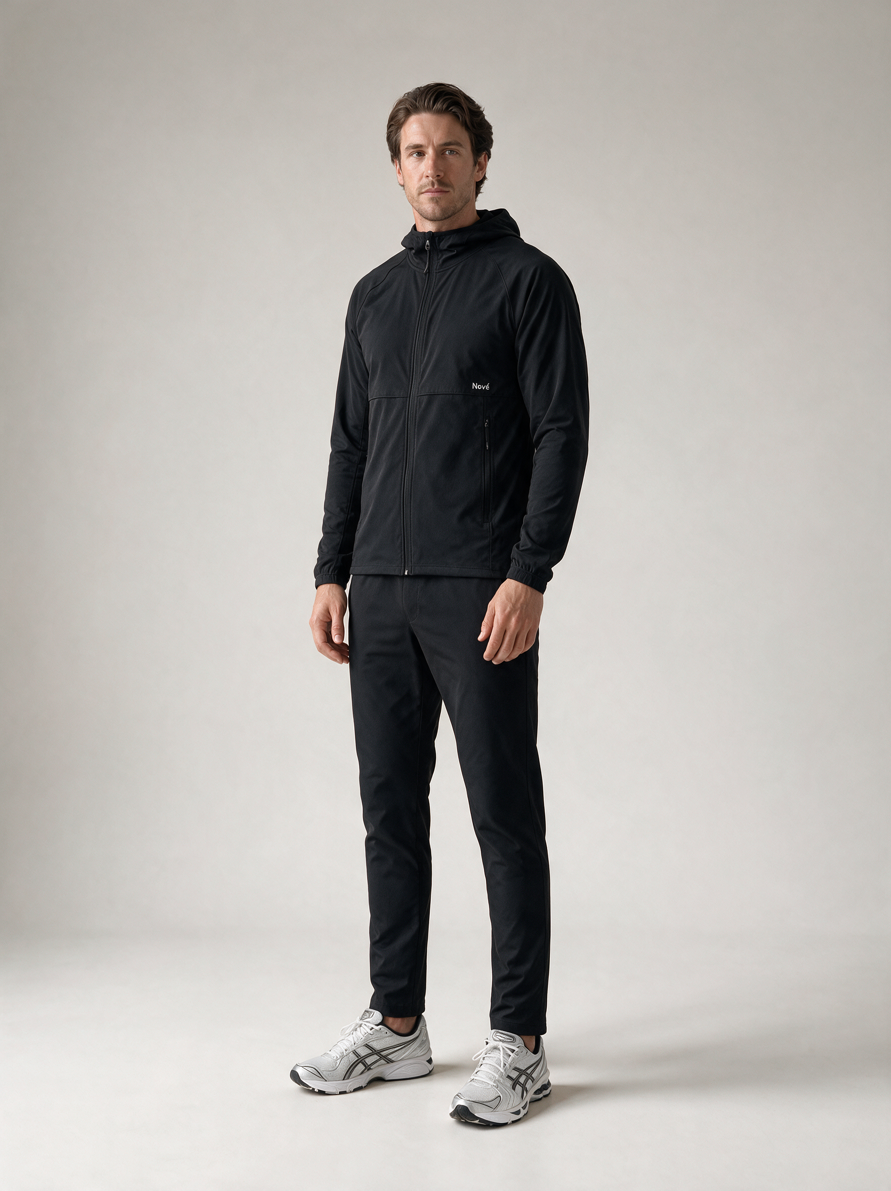 A man modeling a black zip-up hooded jacket and matching black pants with white athletic sneakers, standing against a light gray studio background.