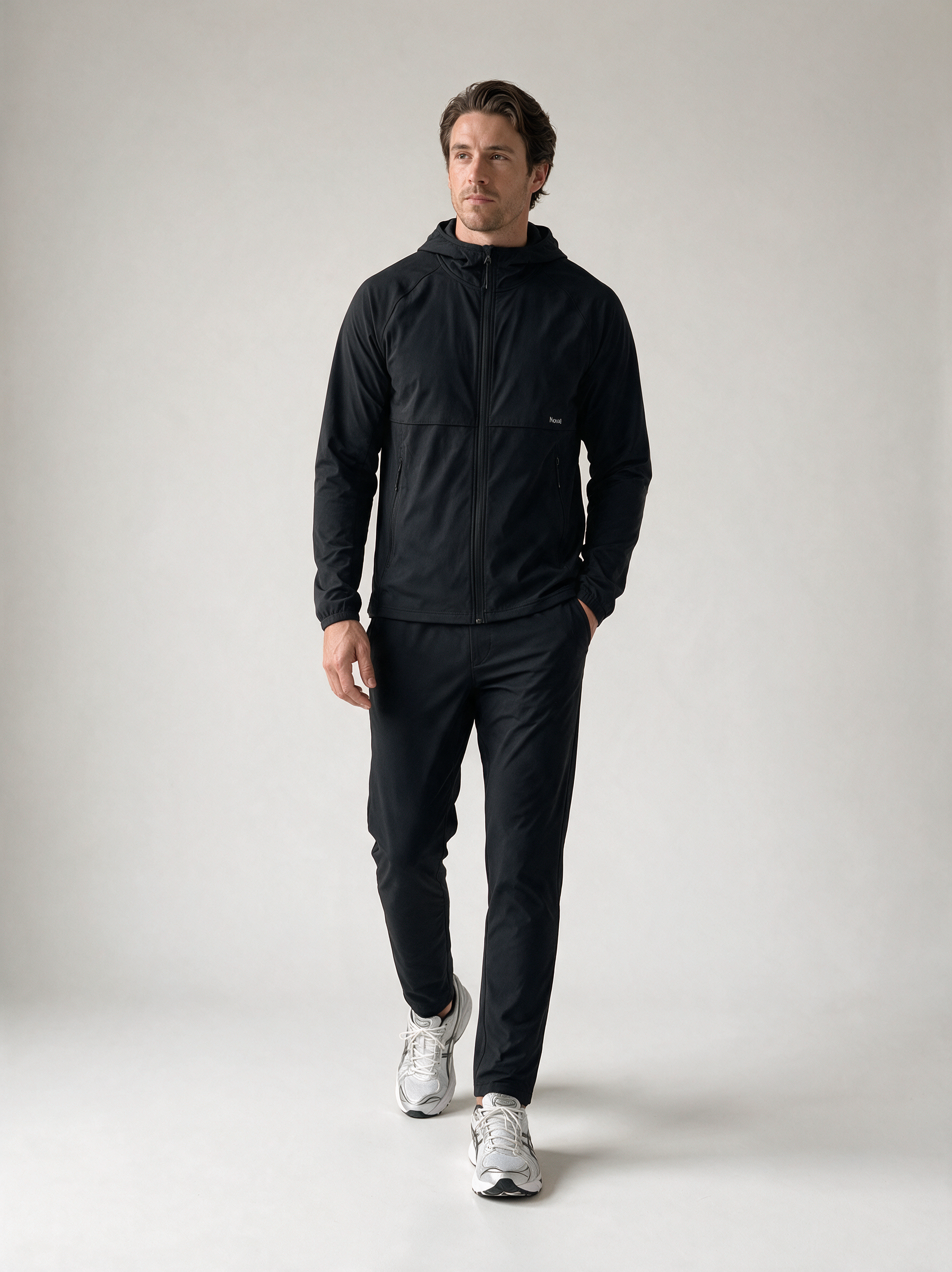 A man wearing a black hooded zip-up jacket and matching black pants, walking forward in a studio setting with white sneakers.