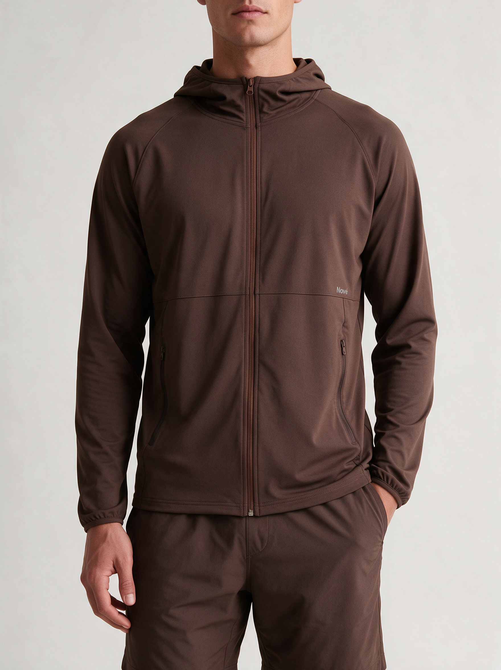 A man wearing a dark brown hooded zip-up jacket and matching brown pants, photographed against a clean light gray studio background.