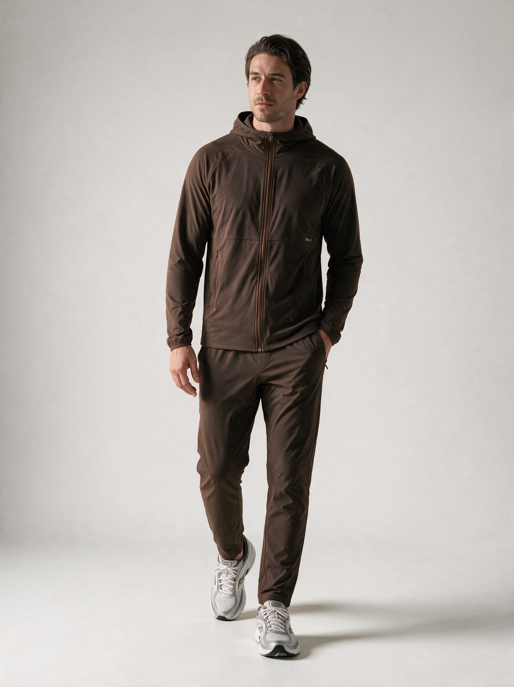 A man wearing a dark brown hooded zip-up jacket and matching dark brown pants, paired with white and gray running shoes, standing against a light gray studio background.