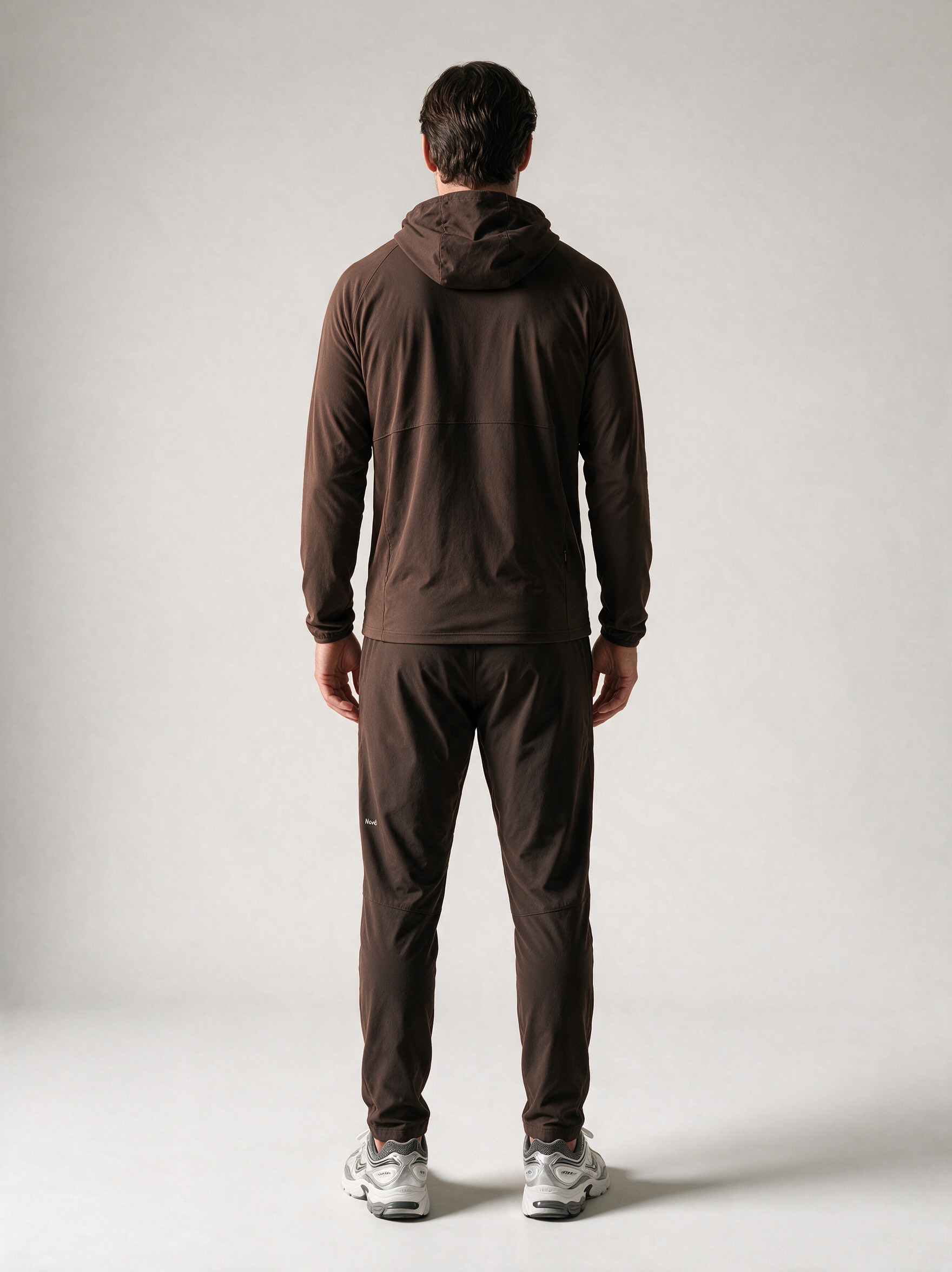 A man viewed from behind wearing a dark brown hooded jacket and matching dark brown pants, standing against a light gray studio background.
