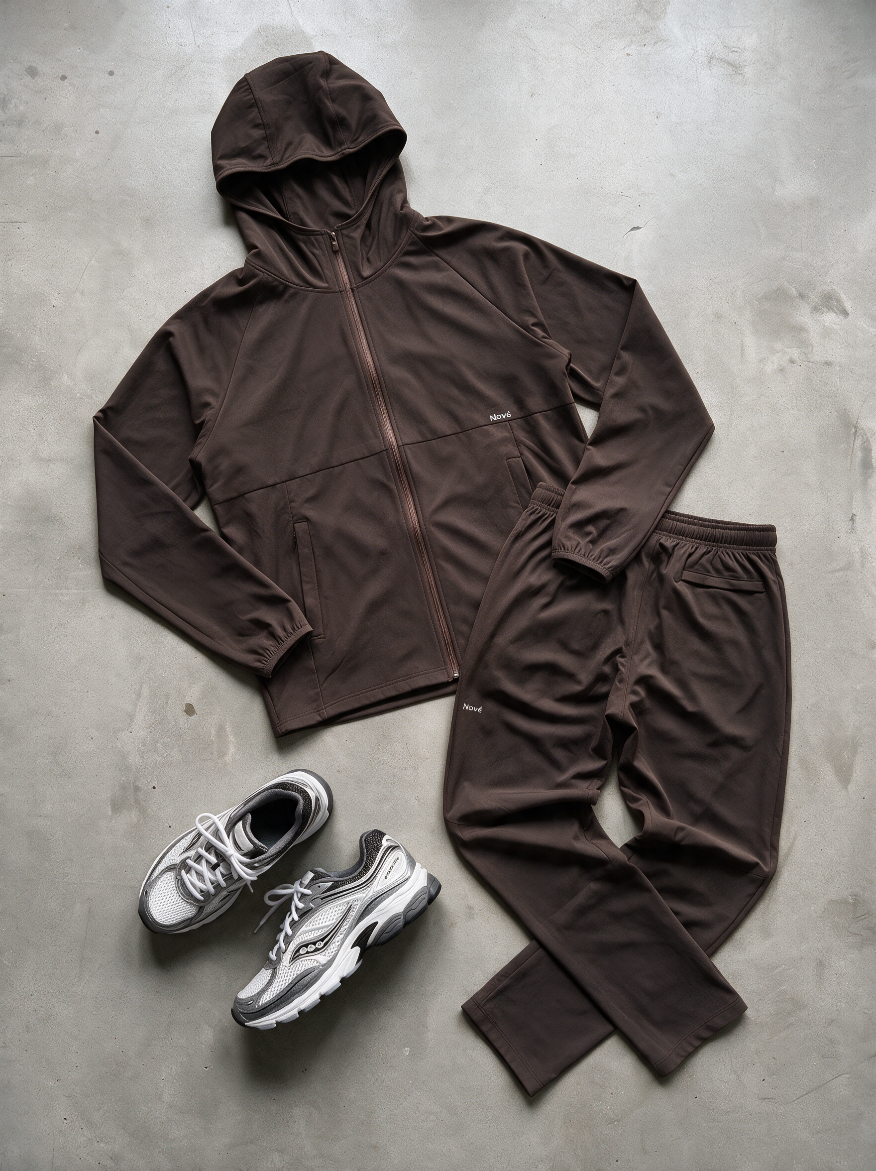 A dark brown athletic tracksuit set consisting of a zip-up hooded jacket and matching sweatpants, laid out flat on a gray concrete floor next to a pair of gray and white running shoes.
