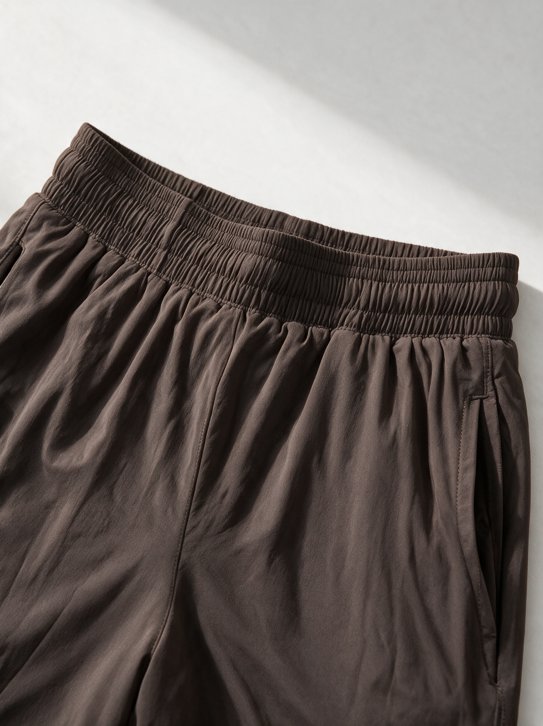 Close-up of dark brown elastic-waist pants with gathered fabric and side pockets, photographed on a light neutral background.