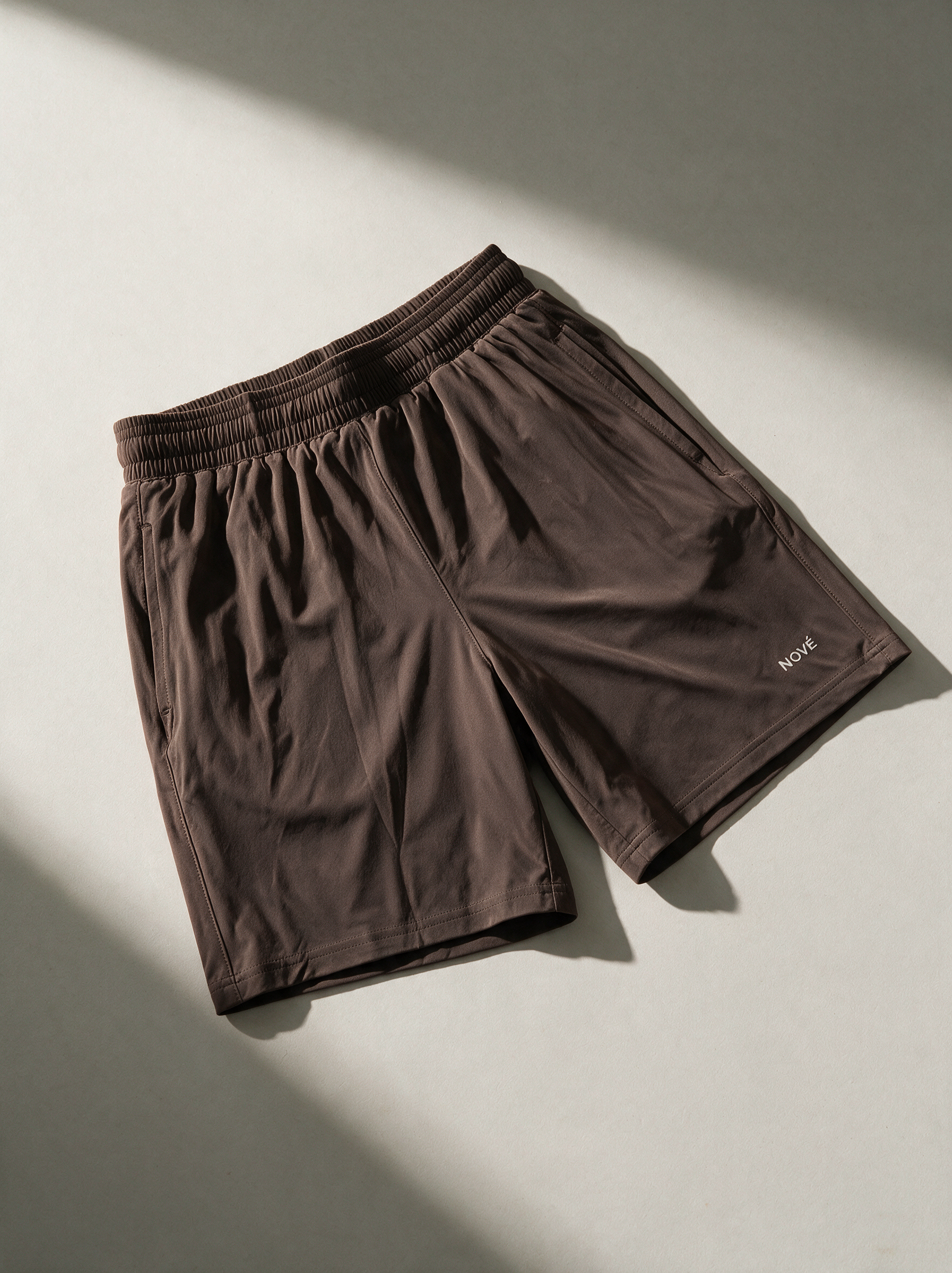 A pair of dark brown athletic shorts with an elastic waistband, laid flat on a light gray surface with diagonal shadows.